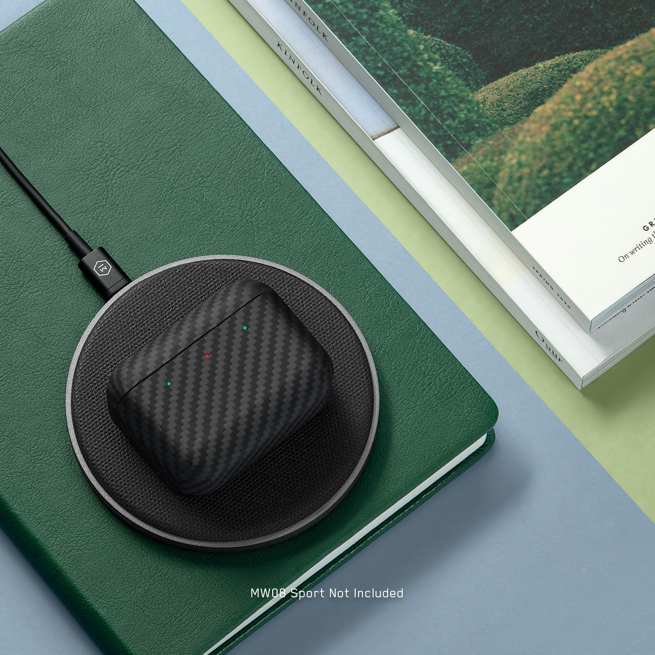 A gunmetal MC100 Wireless Charge Pad on a table with books and green stationary, charging a closed black Kevlar earphone case | gallery MC100G1