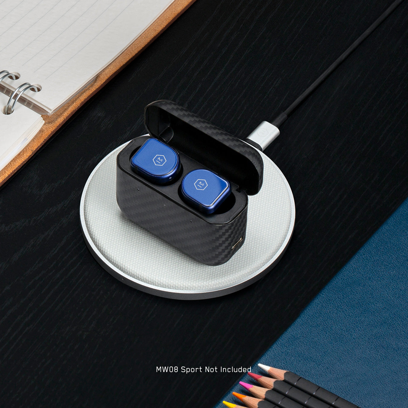 A silver MC100 Wireless Charge Pad on a black wooden table with stationary and pencils underneath a pair of blue earphones inside their case | gallery MC100S3