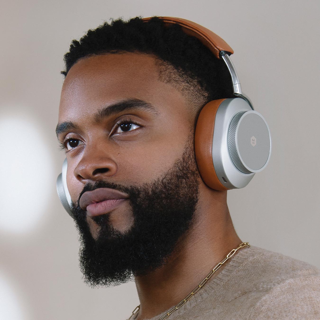 Close up of a bearded man wearing a tan sweater and gold chain wearing silver and brown MW75 Active Noise-Cancelling Headphones against a beige background | gallery MW75S2